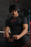 A muscular man with a mustache sits in a gym wearing a tight black t-shirt with a red logo on the left chest. He has large over-ear headphones on his head and is looking down at a shaker bottle or object held in his hands.