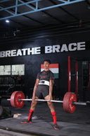 A front view of a person wearing a lifting belt and red socks performing a sumo deadlift with a heavily loaded barbell inside a gym. On the black wall in the background, bold white text reads "BREATHE BRACE," and an Indian flag is visible to the right.